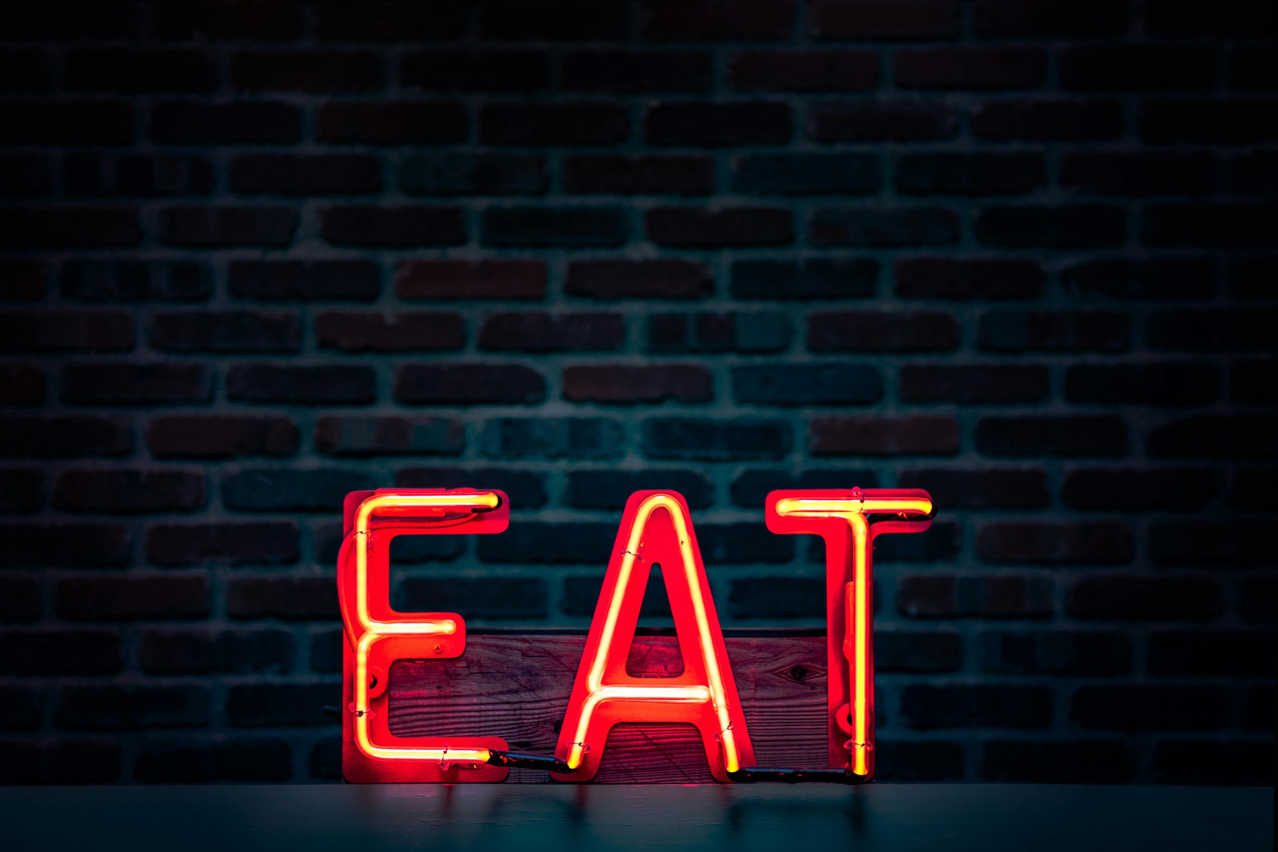 Vibrant neon 'EAT' sign in red glowing boldly against a dark brick wall background.