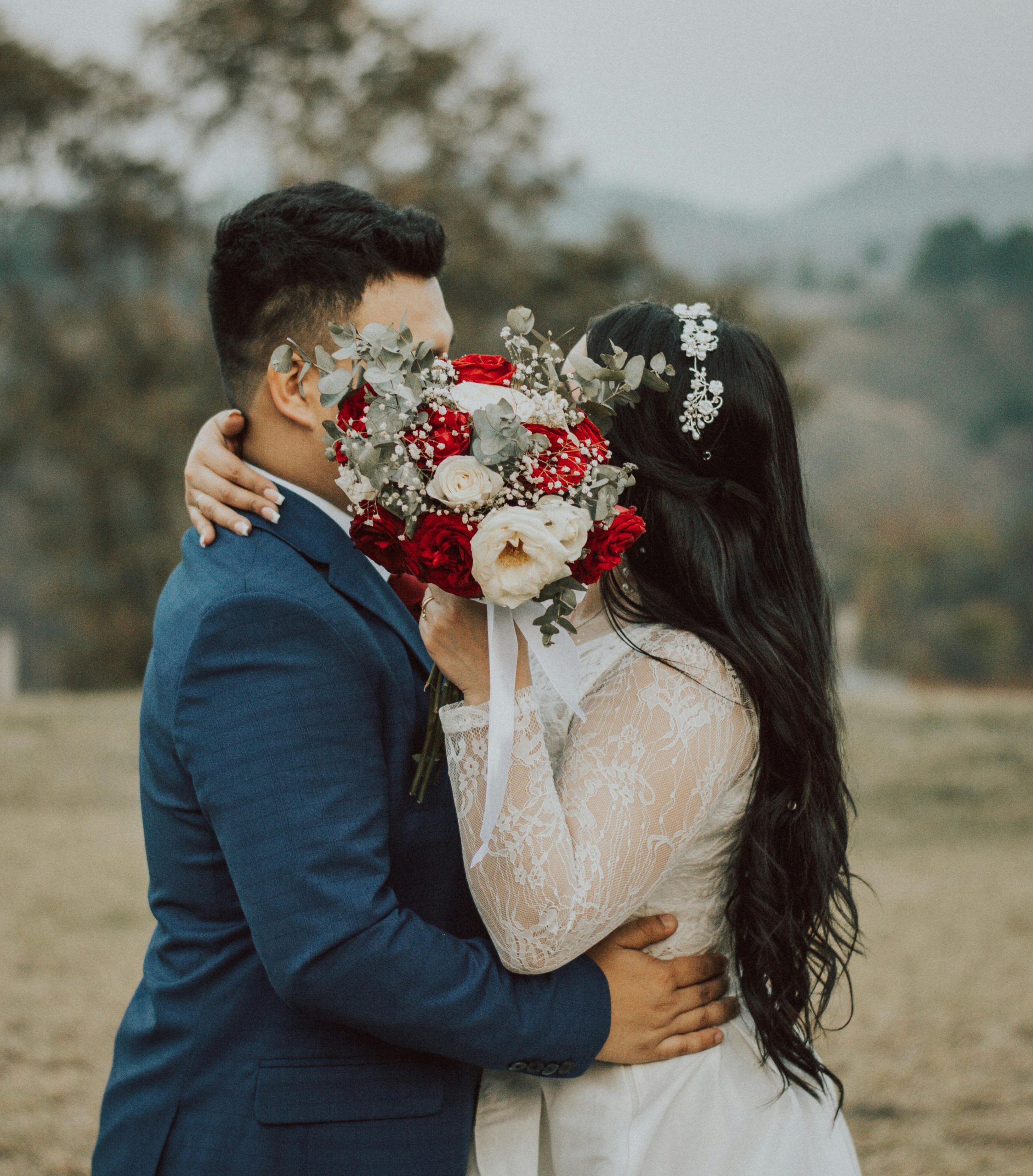 Bride and groom share a hidden kiss behind a bouquet in an outdoor setting.