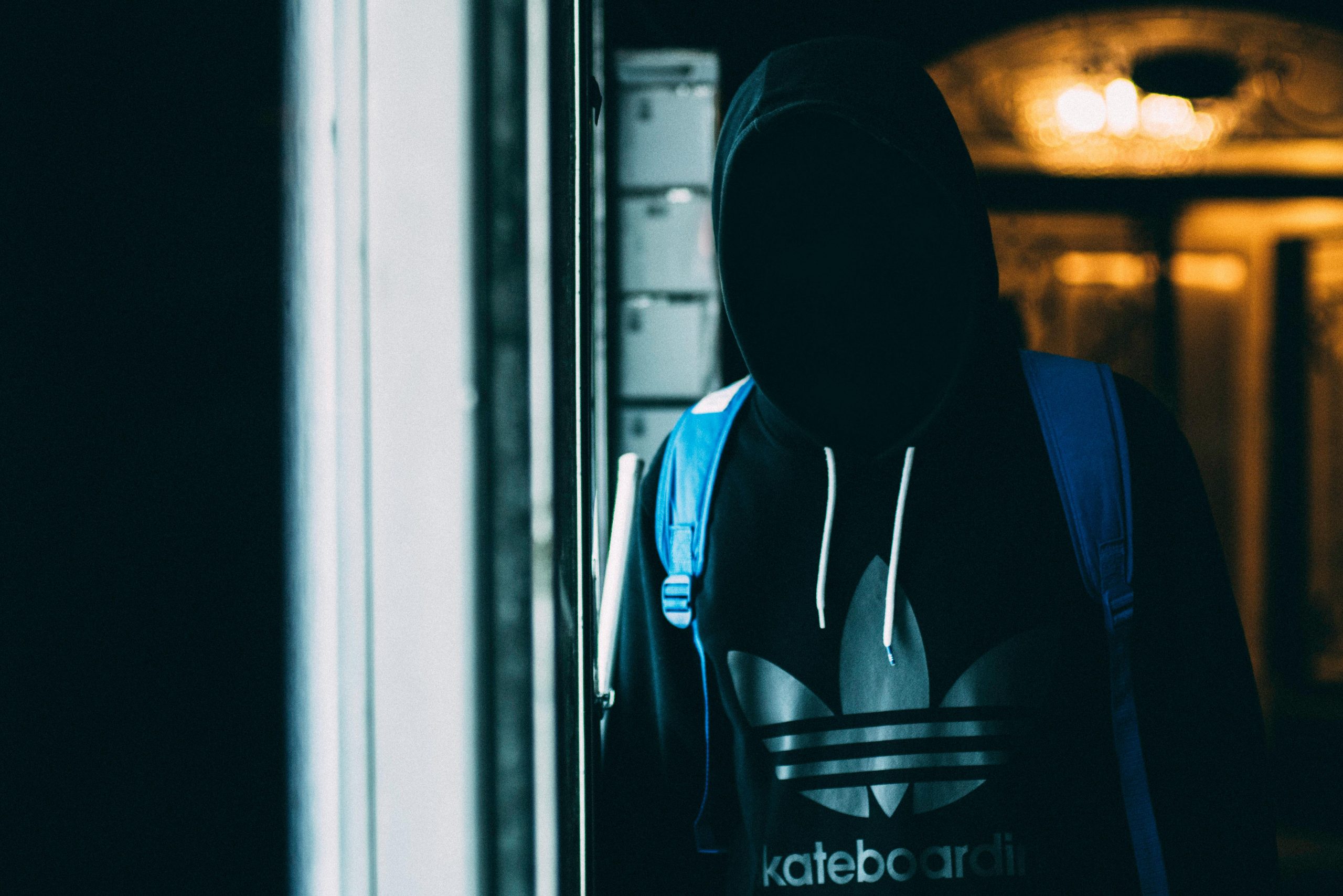 A shadowy figure in a hoodie stands with a backpack in dim lighting, conveying mystery and anonymity.