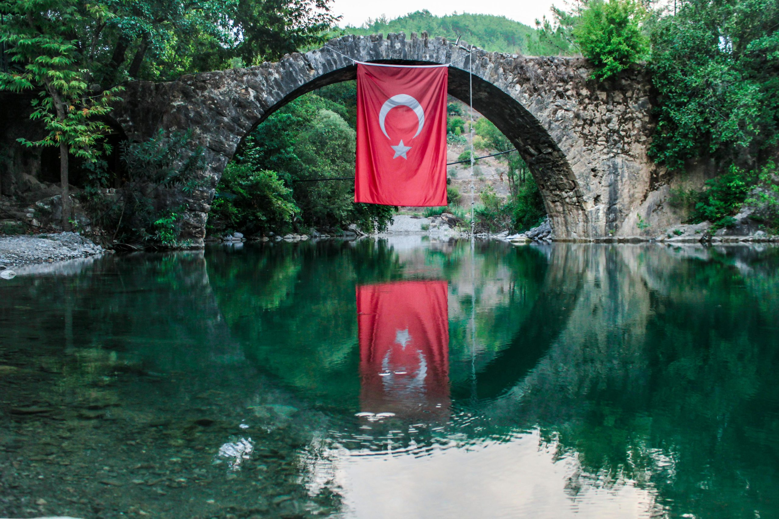 A serene view of a stone bridge with a Turkish flag reflecting on the water below.