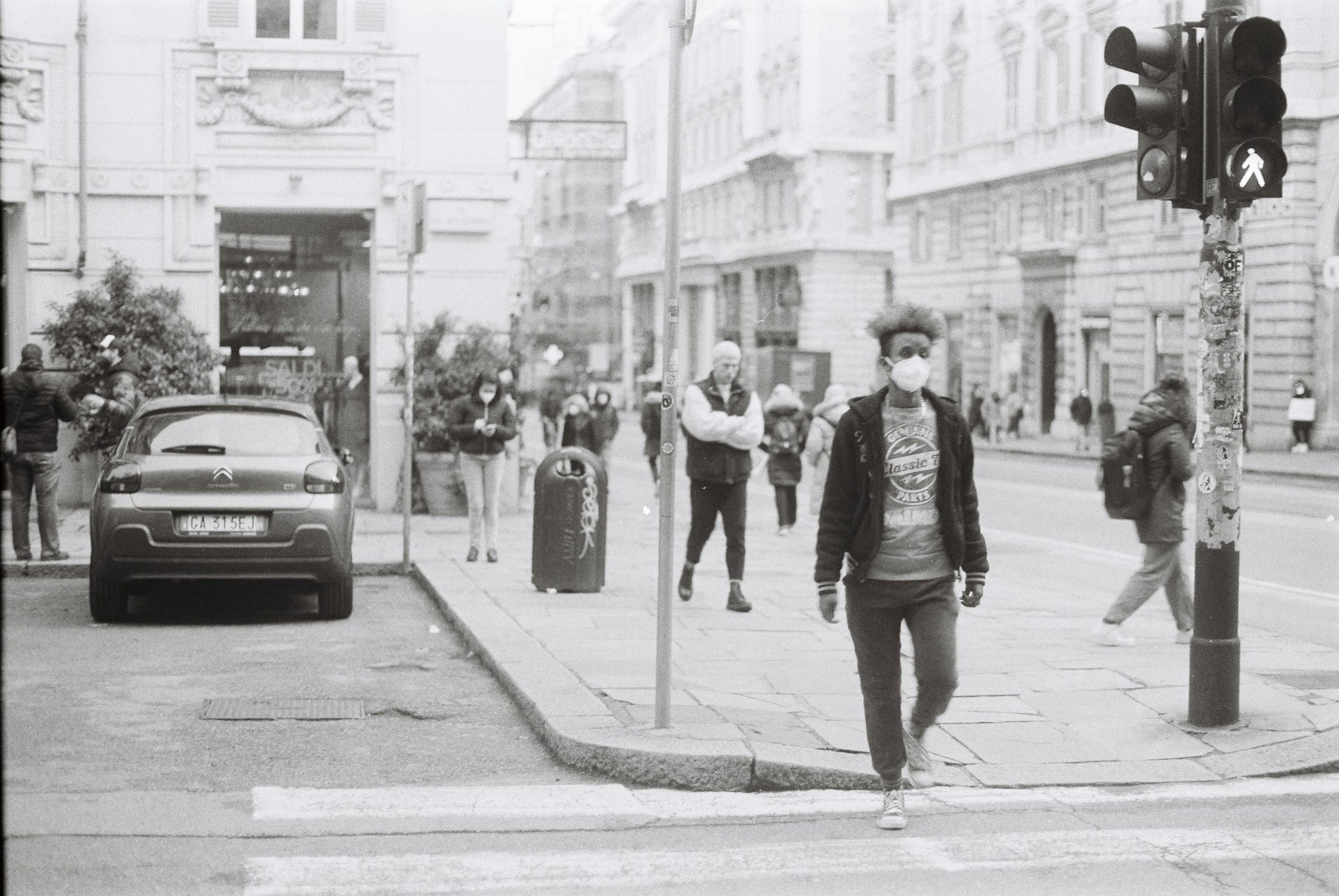 Black and white street scene with pedestrian wearing mask in urban setting.