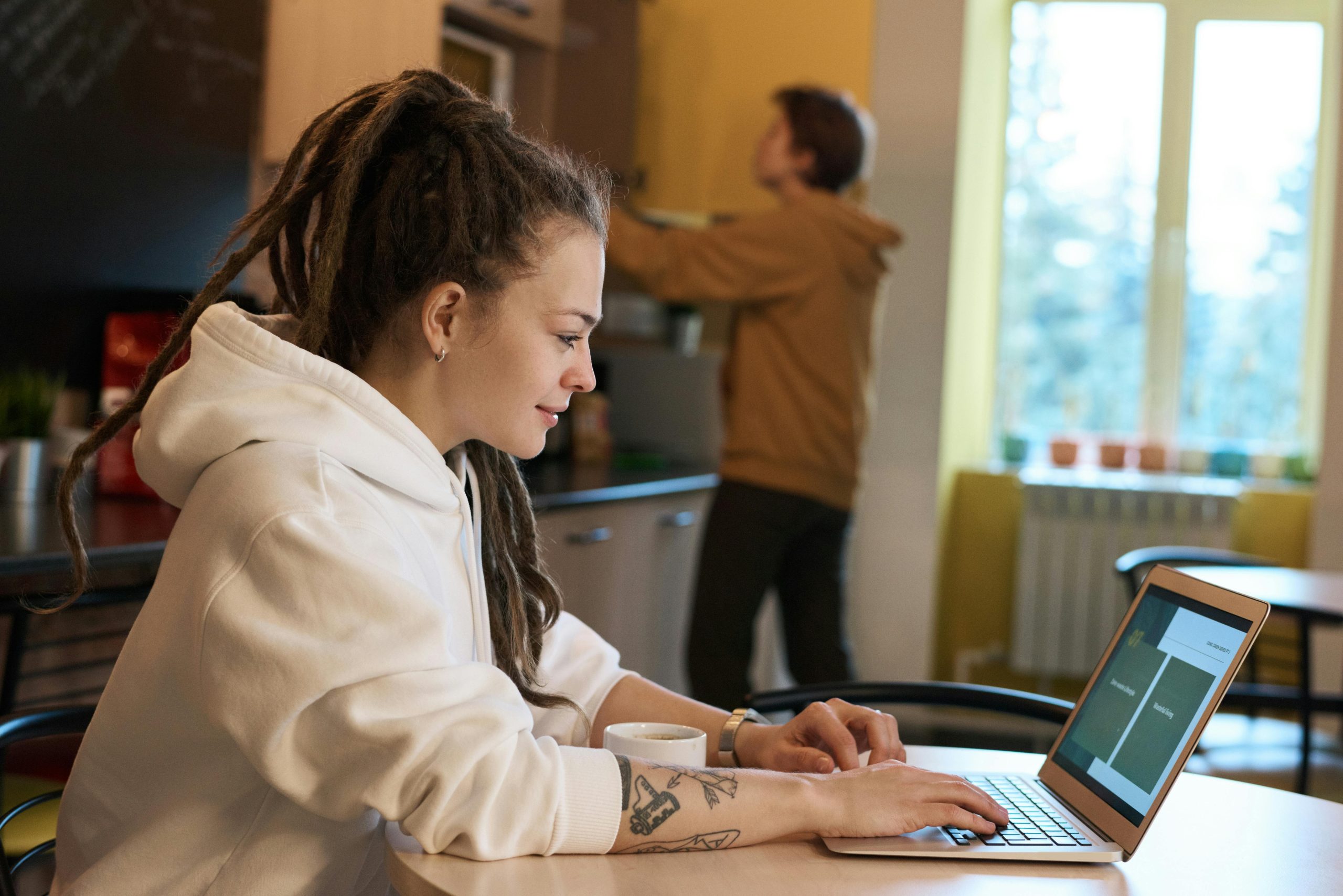 Young woman with dreadlocks works on a laptop in a casual kitchen environment.