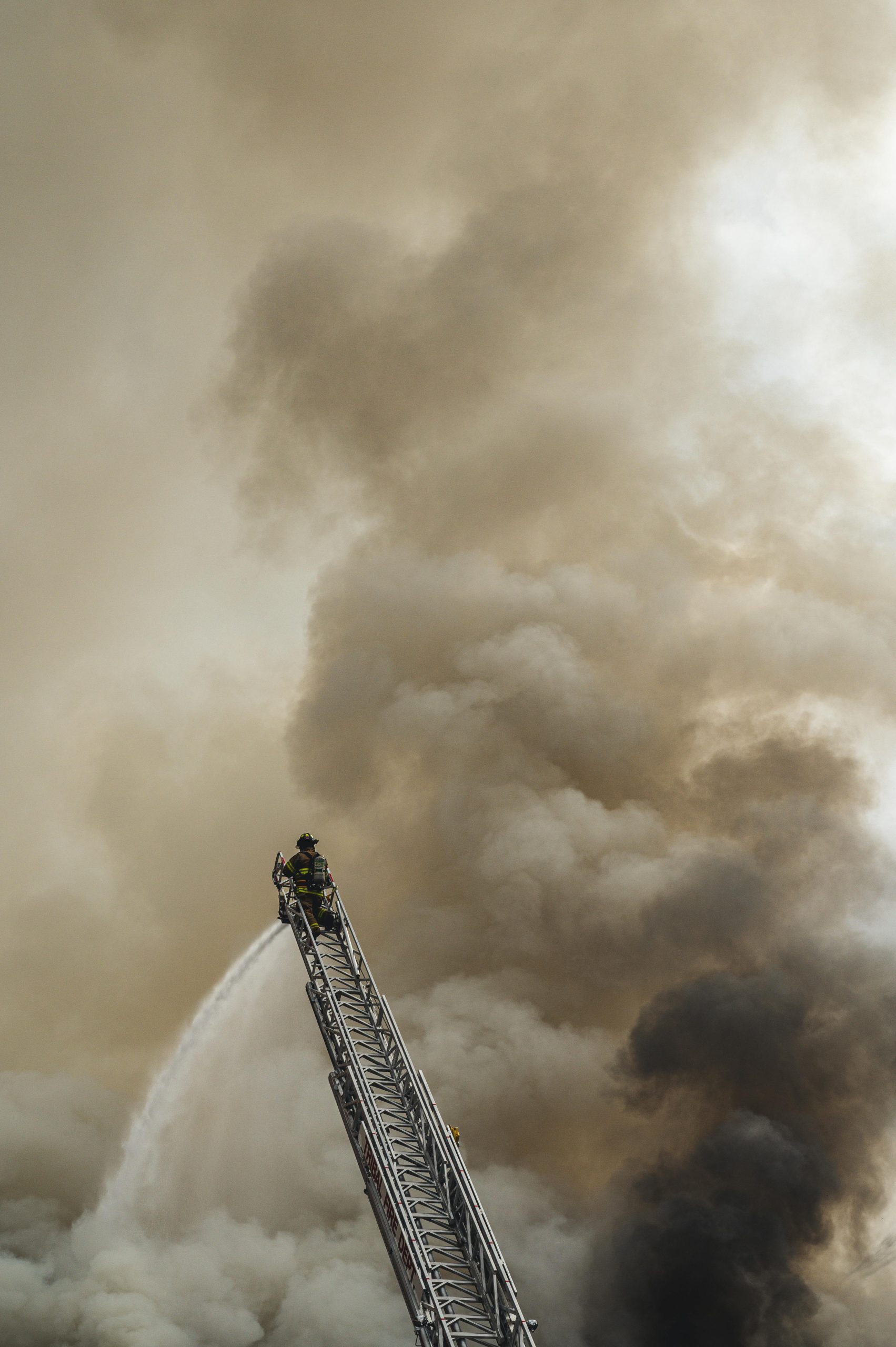 Brave firefighter extinguishing fire from a ladder amidst heavy smoke backdrop.