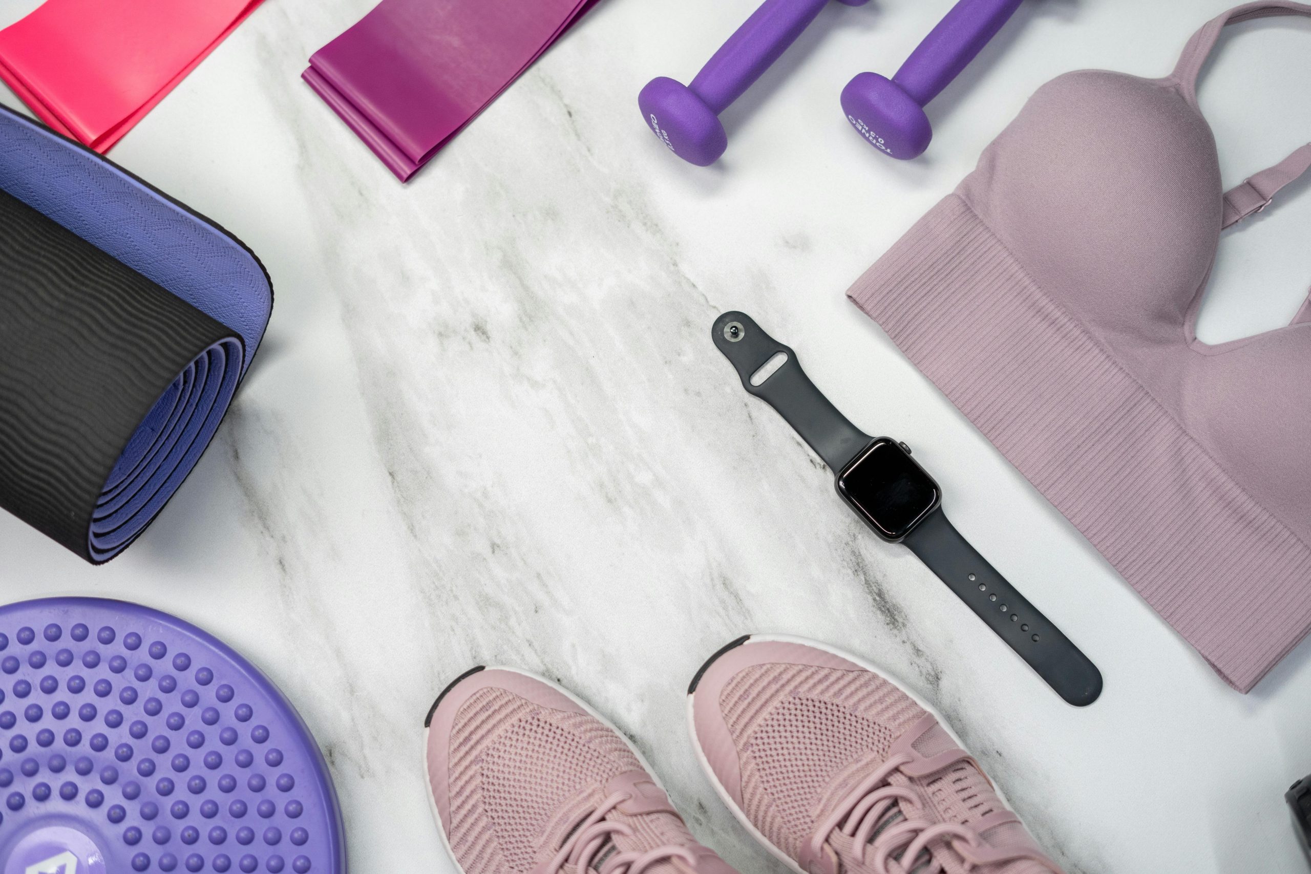 Flat lay of fitness gear including yoga mat, dumbbells, and smartwatch on a marble surface.