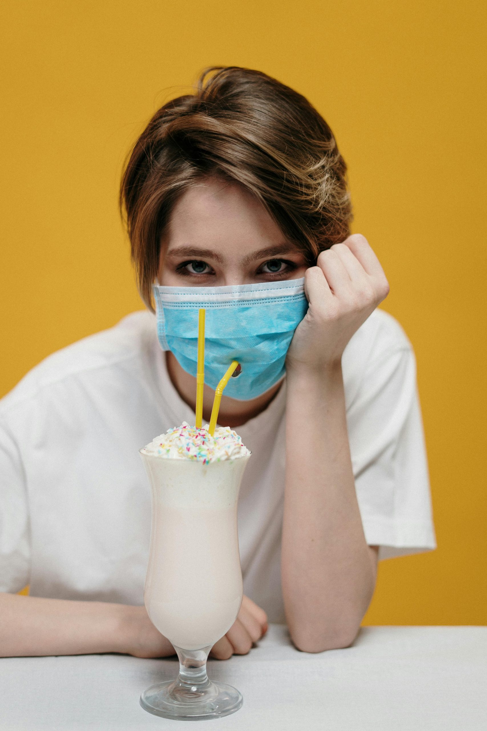 A woman in a face mask with a milkshake in a humorous portrait, highlighting pandemic lifestyle.