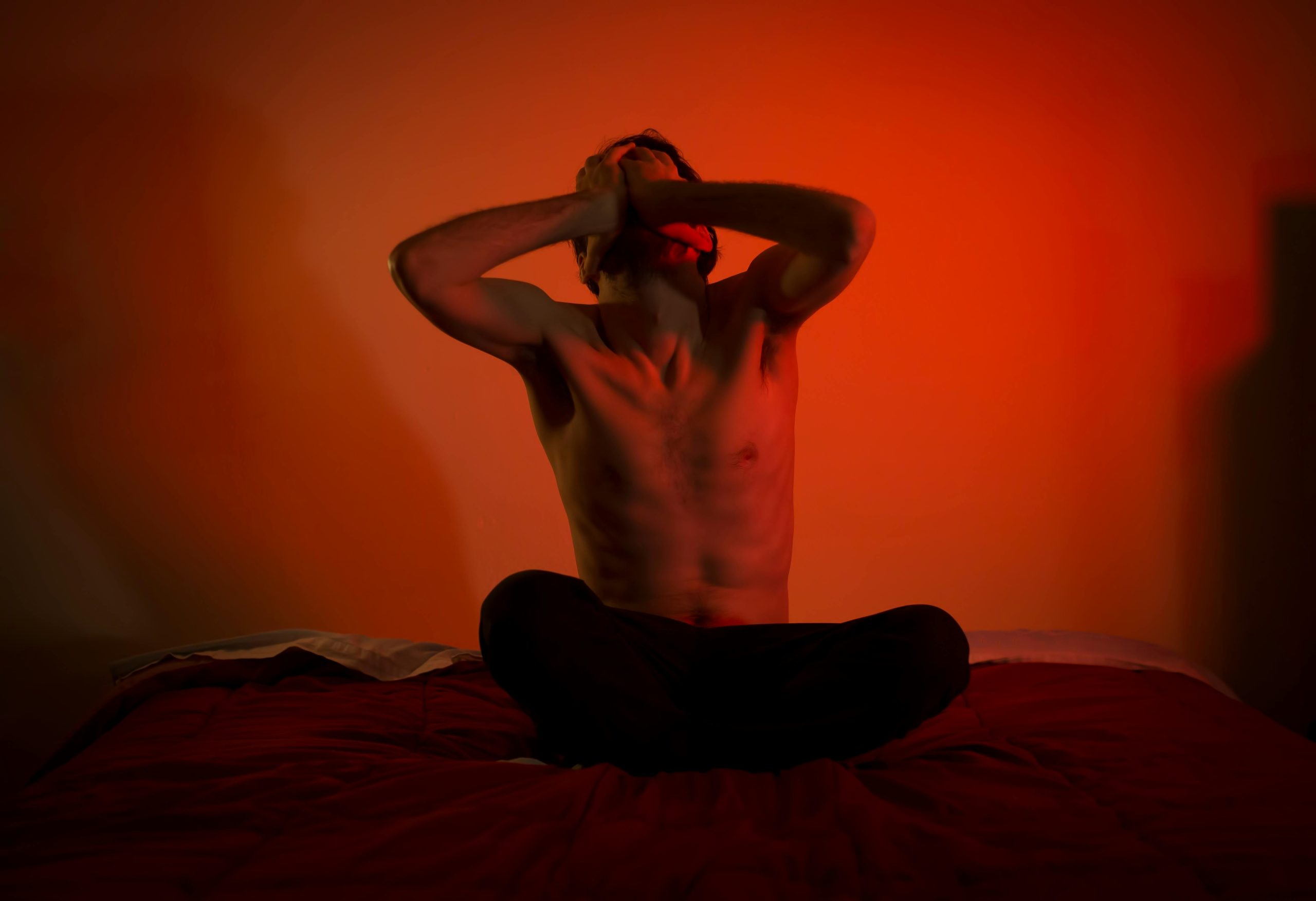 A topless man showing emotional distress sitting on a bed in a dimly lit room.