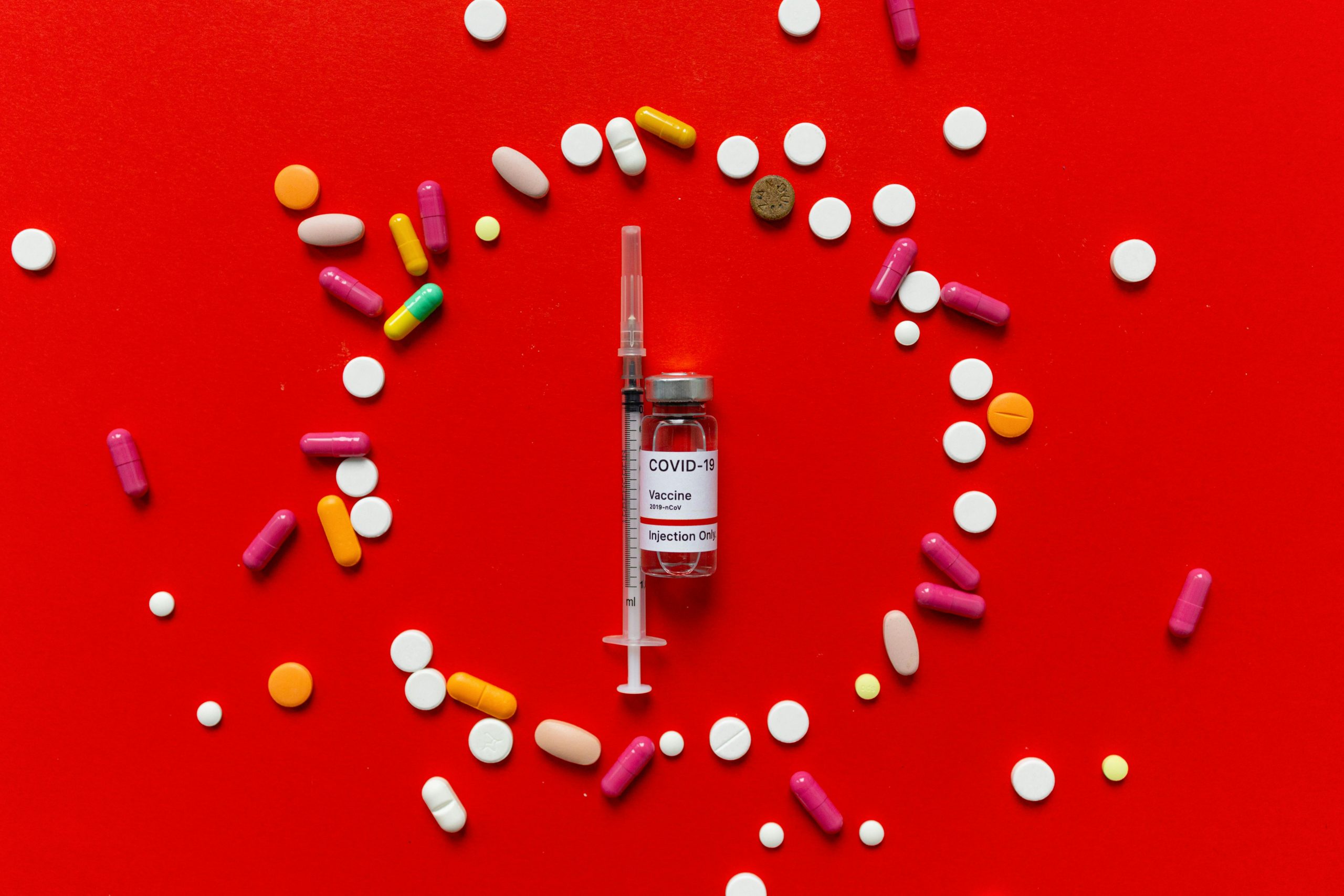 Vaccine vial and syringe surrounded by colorful pills on vivid red surface.