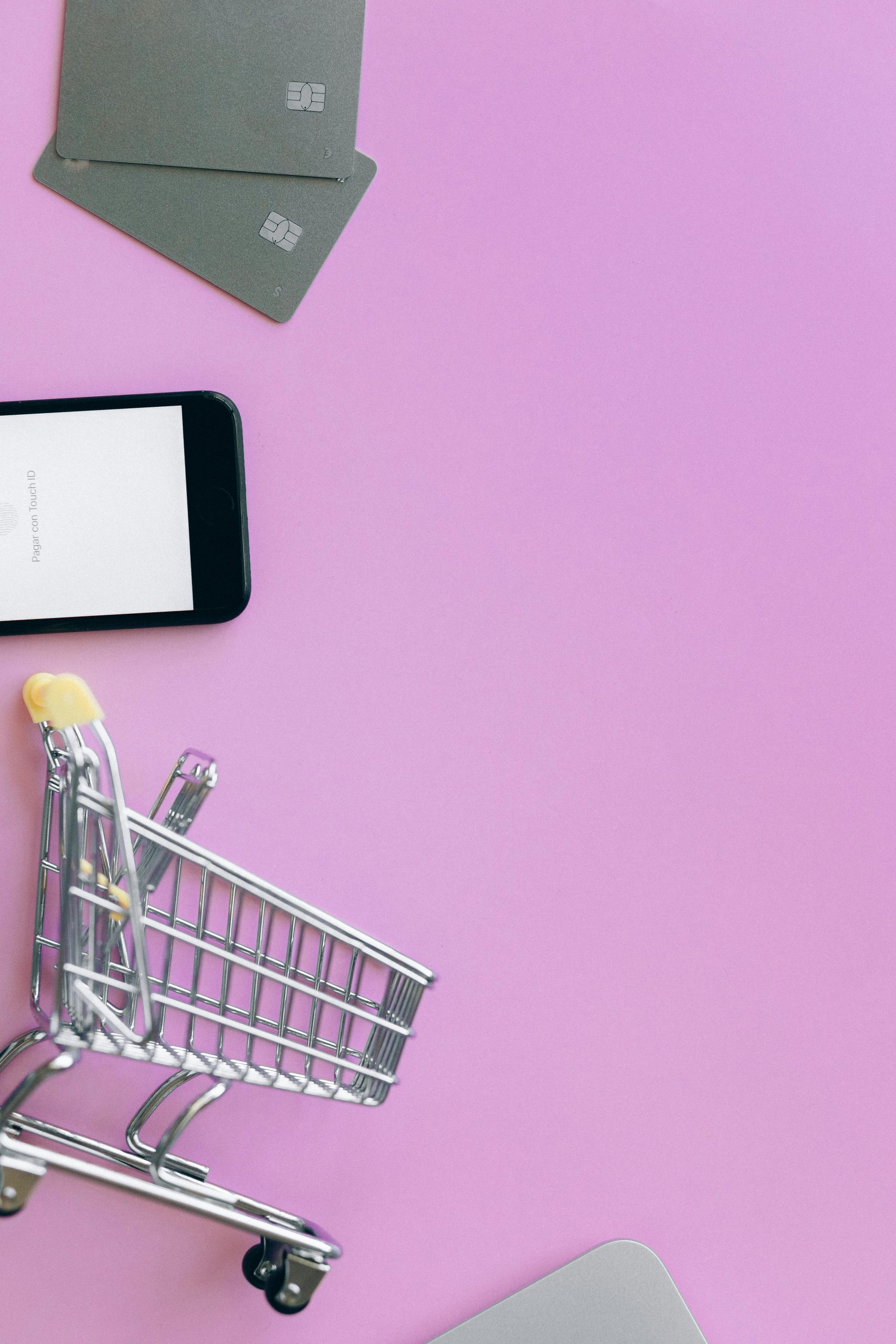 Top view of miniature shopping cart with credit cards and smartphone on pink background.