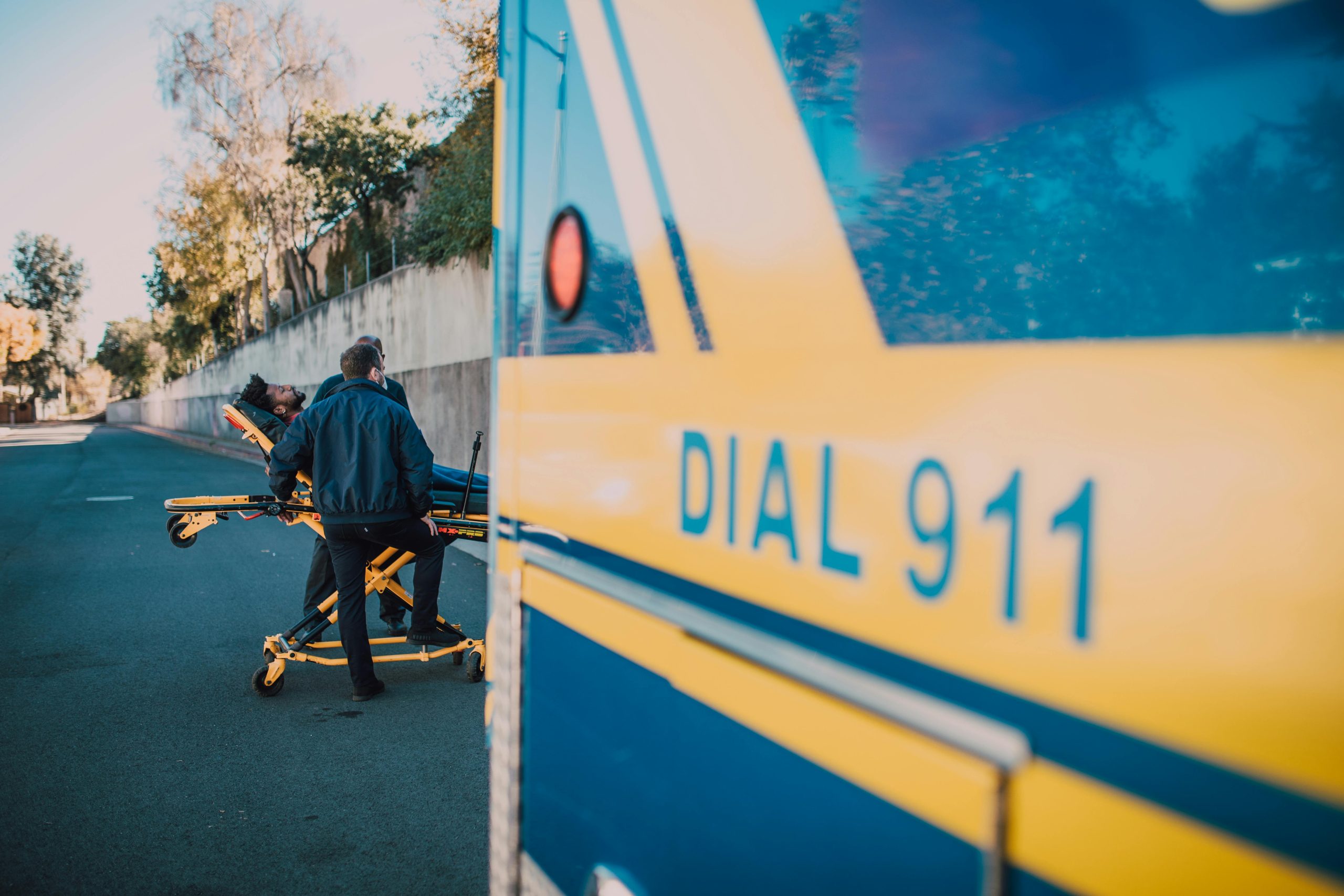 Paramedics assist a patient on a stretcher near an ambulance labeled 'Dial 911' on a sunny day.