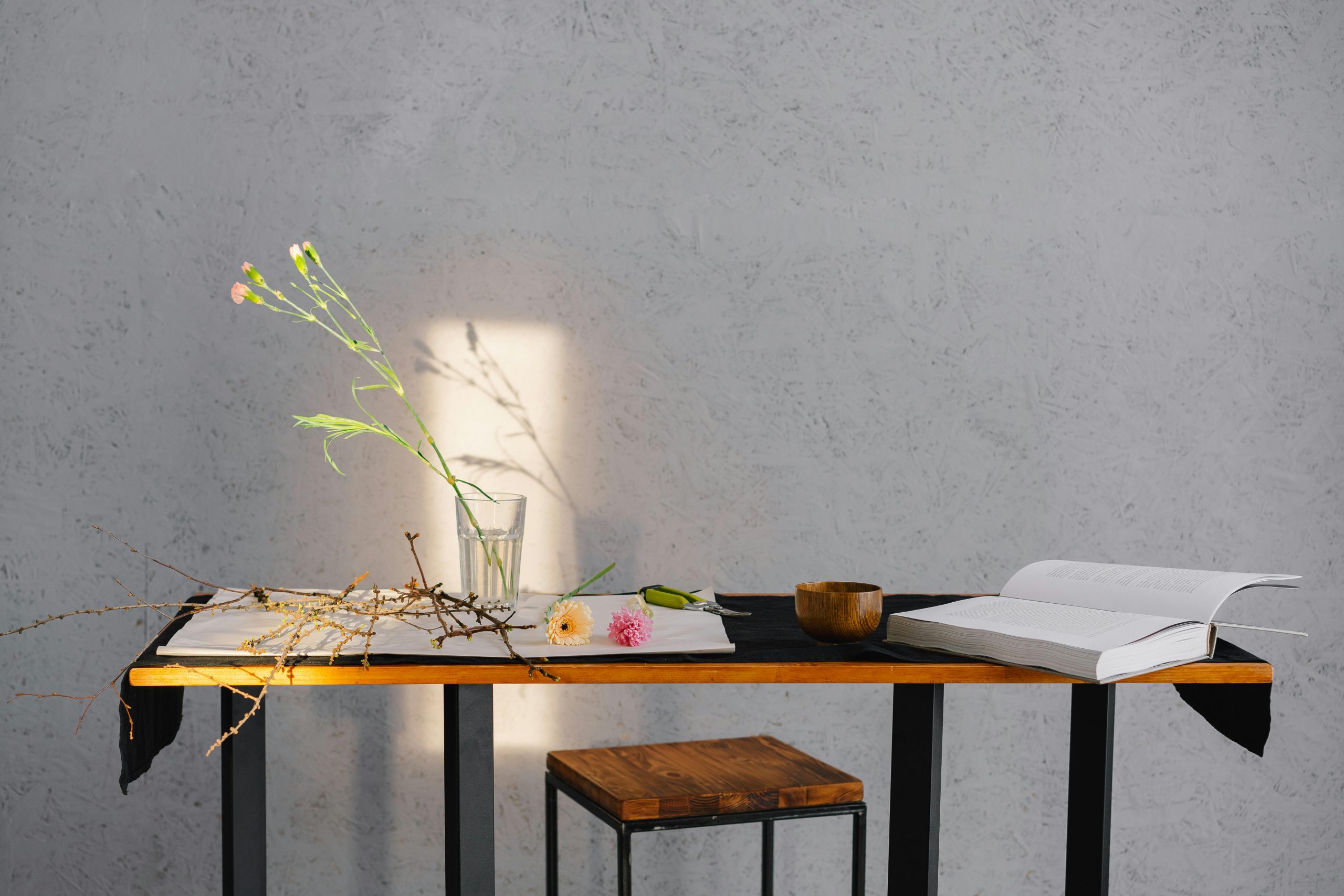 A minimalist scene of a table with flowers, an open book, and wooden bowl, enhanced by natural lighting.