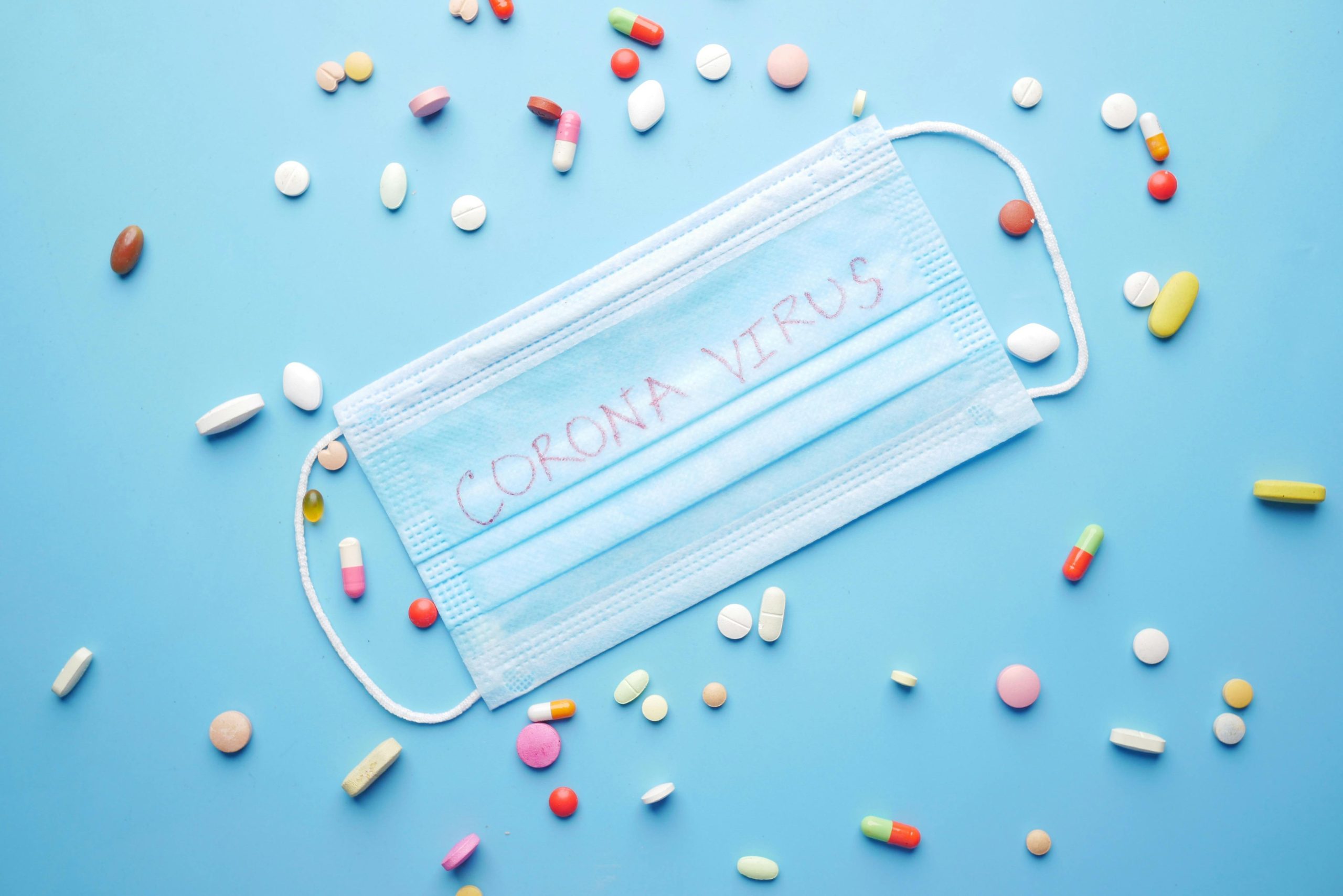 Top view of mask labeled 'corona virus' surrounded by pills on a blue background.