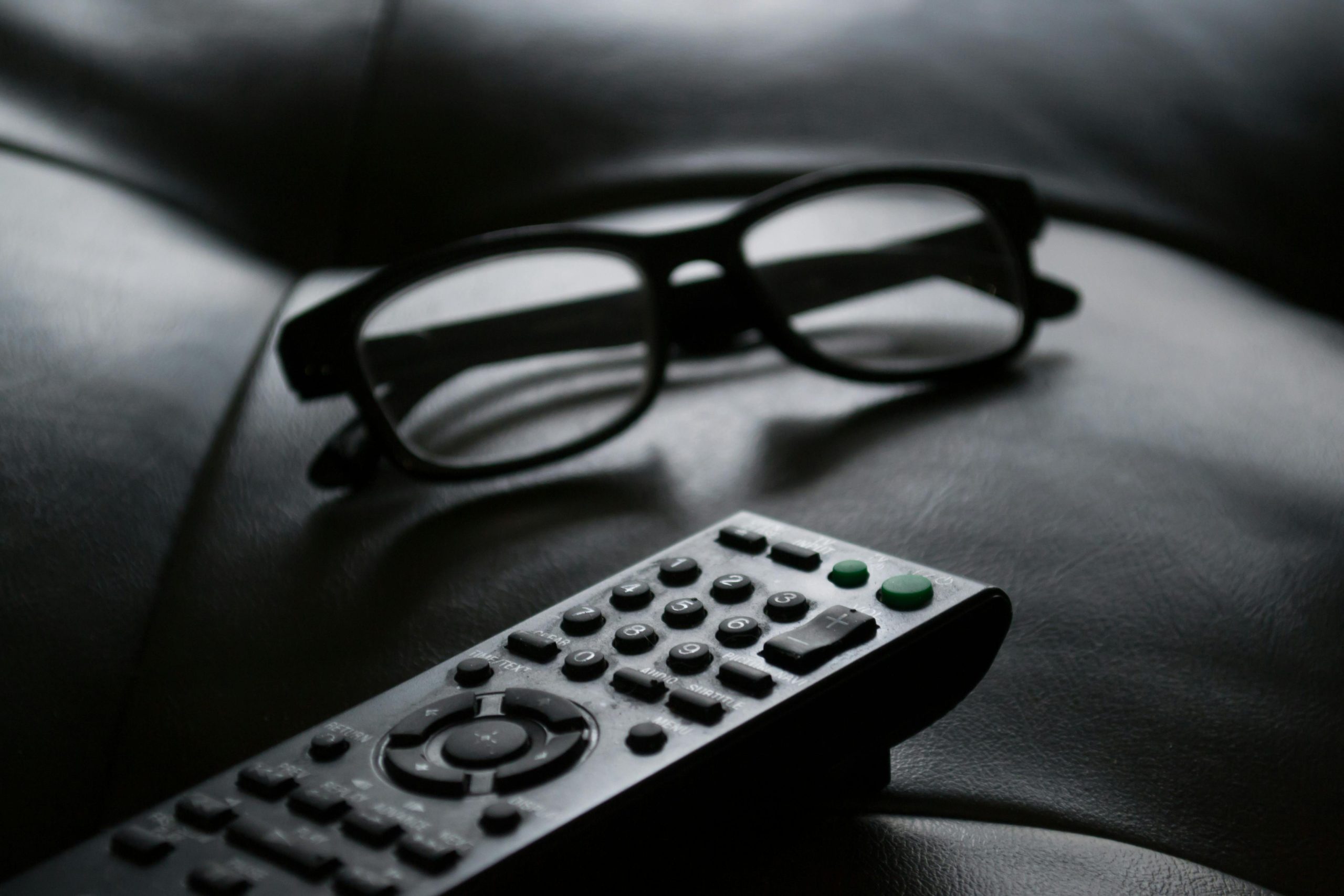 Close-up of eyewear and a TV remote on a dark leather couch, ideal for home lifestyle themes.